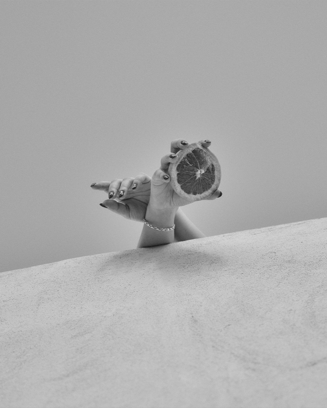 A black and white photo of a hand holding a slice of grapefruit. The hand is emerging from behind a textured surface, and the person is wearing a ring and bracelet.